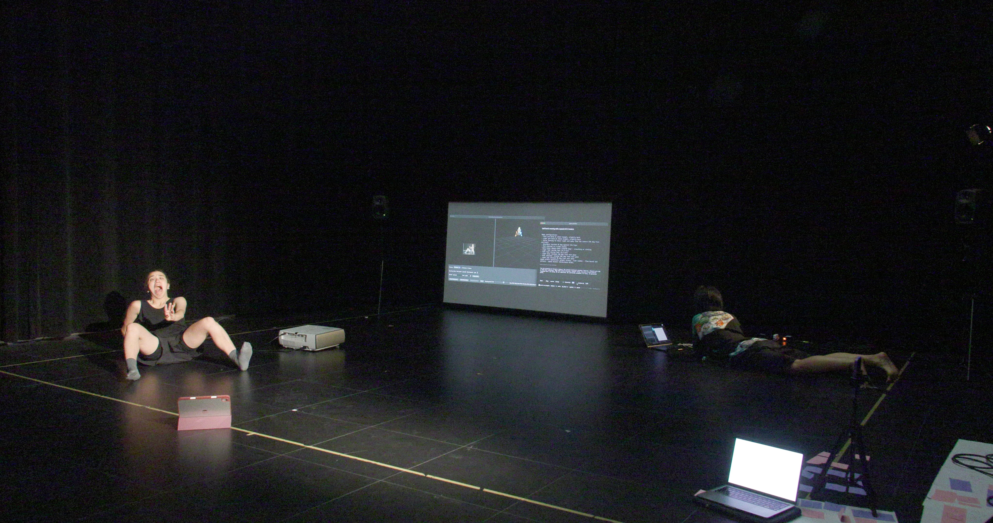 a black box space with a large projection on the back and one female dancer siting on the floor to the right and one female performer working on her laptop on the floor to the left